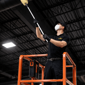 Technician performing high dusting from a scissor lift to remove overhead dust in a warehouse.
